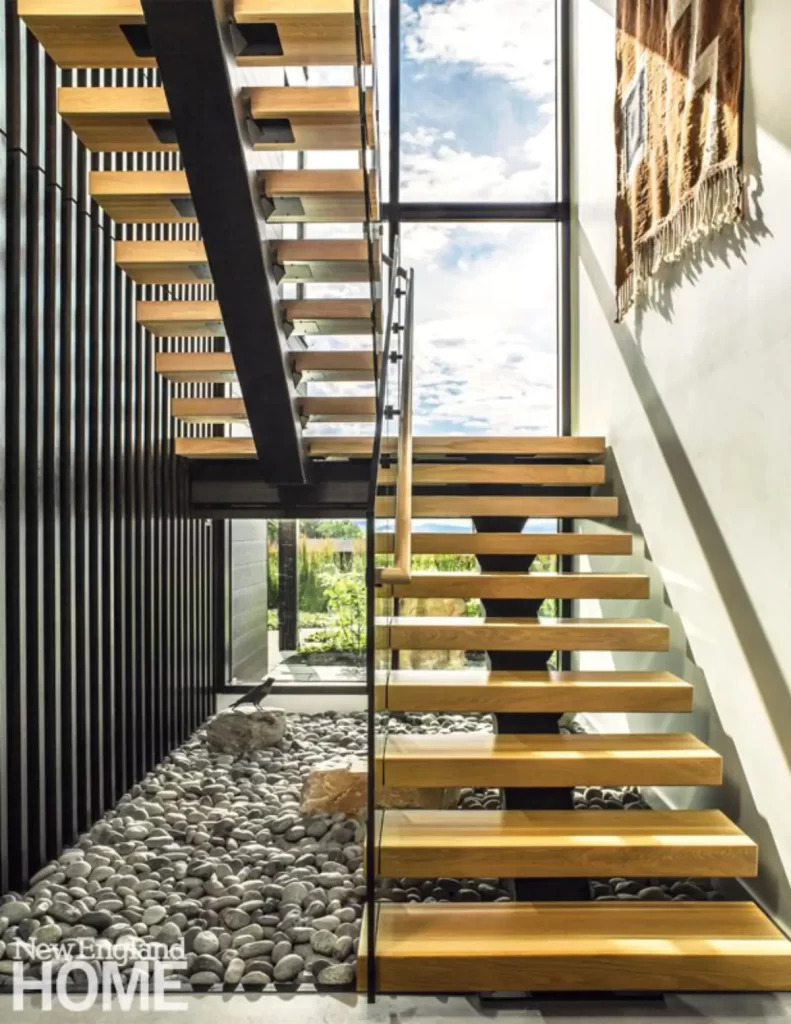 راه پله ای مدرن از چوب با سنگ های رودخانه ای در کف و نمای شیشه ای بلند که به ارتباط بصری بیرون وداخل کمک میکند